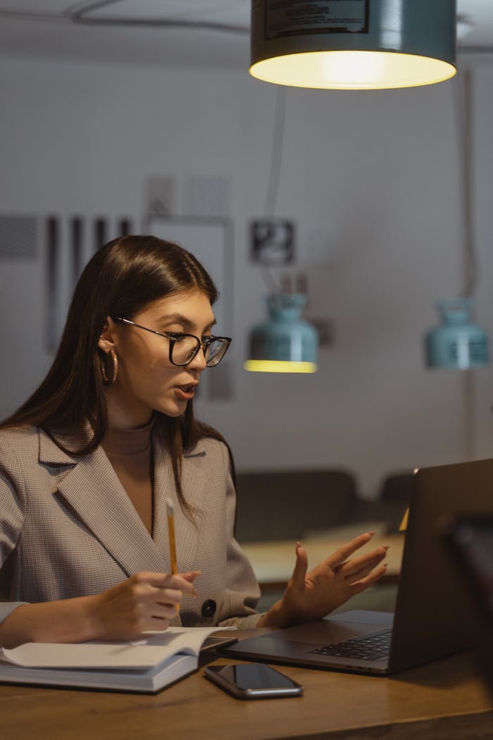 A young businesswoman working late at her desk, engaged in an online meeting with a laptop, notebook, and smartphone.